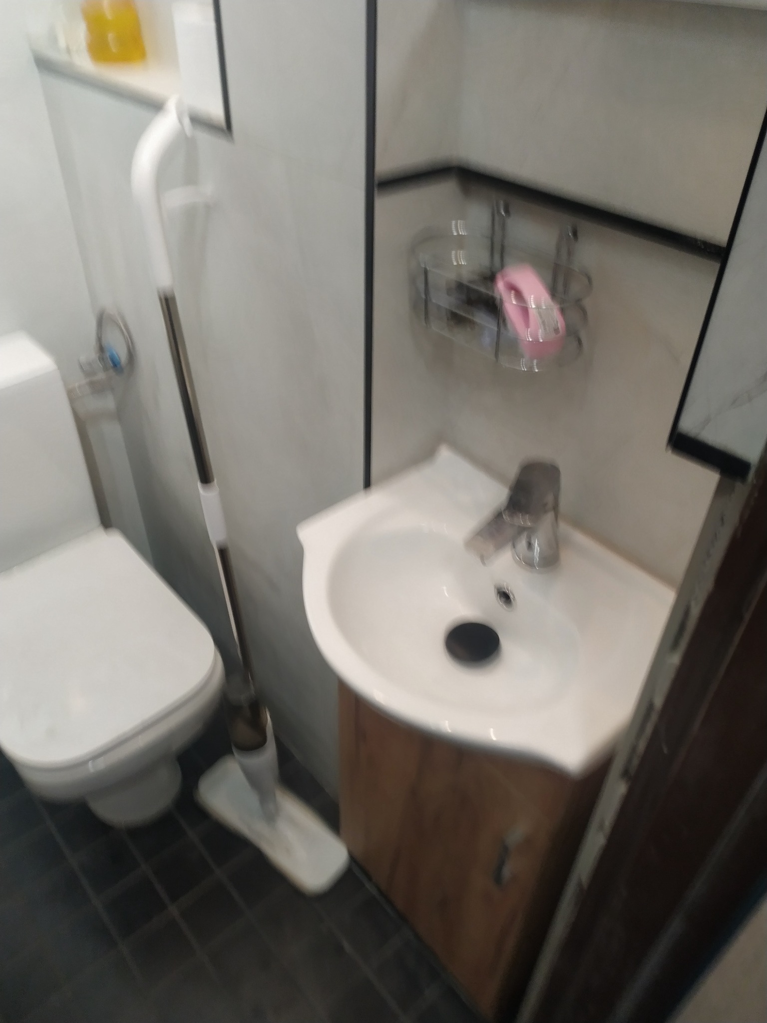 Umywalka narożna z szafką w małej łazience, obok toaleta, na ścianie półka z akcesoriami, widoczny mop stojący na podłodze.