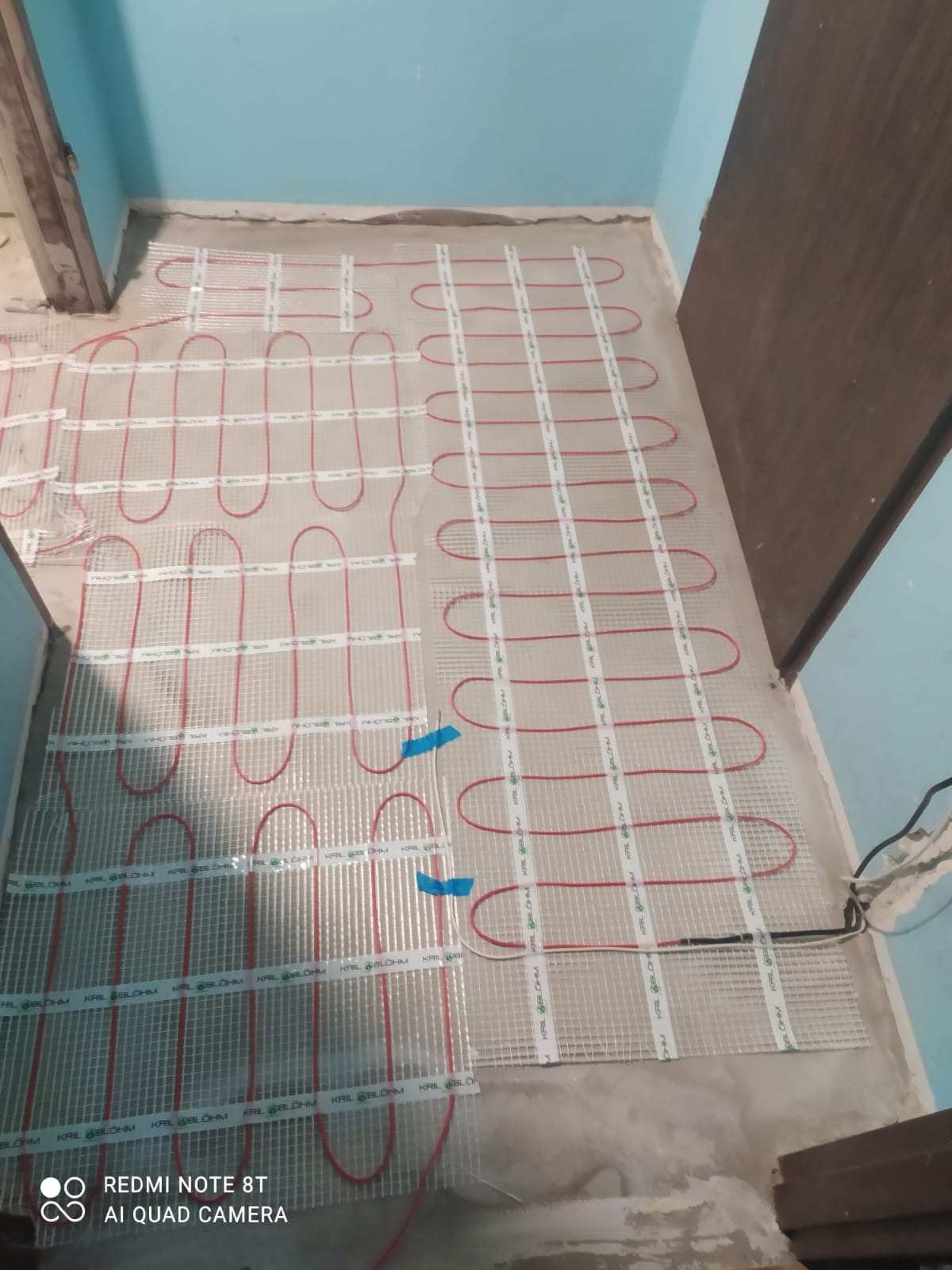Rozłożona mata grzewcza na podłodze w trakcie montażu, widoczne czerwone kable grzewcze ułożone w pętle na siatce, przytwierdzone niebieską taśmą, przygotowanie do wylewki.