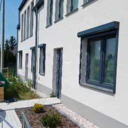 Elewacja budynku z zamontowanymi roletami zewnętrznymi w kolorze antracytowym. Widoczny fragment drabiny i rabaty z korą.