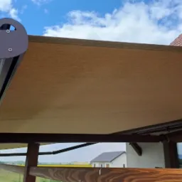 Beżowa markiza tarasowa z falbaną, zamontowana przy drewnianej konstrukcji. W tle widoczne pole rzepaku, dom i błękitne niebo z chmurami.