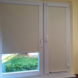 Volodymyr Vorobets - Beżowe rolety zwijane w oknach PCV, jedna częściowo opuszczona, z widokiem na zieleń. Obok firanka i fragment blatu kuchennego z dekoracjami.