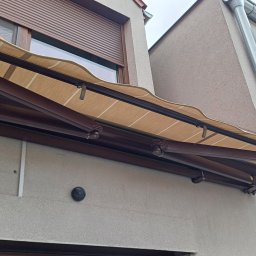 Volodymyr Vorobets - Beżowa markiza tarasowa z brązową konstrukcją zamontowana nad oknem z roletą zewnętrzną. Widoczny fragment rynny i klimatyzator na elewacji budynku.