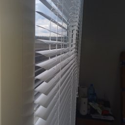 Volodymyr Vorobets - Białe żaluzje poziome, częściowo otwarte, w oknie. Widok z boku, z widocznym fragmentem pokoju i okna z widokiem na budynki w oddali.