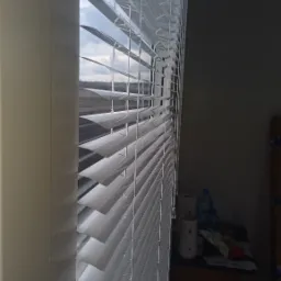 Białe żaluzje poziome, częściowo otwarte, w oknie. Widok z boku, z widocznym fragmentem pokoju i okna z widokiem na budynki w oddali.