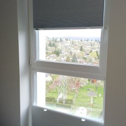 Volodymyr Vorobets - Szara, podwójna plisa okienna w Świdnicy, zamontowana w oknie z widokiem na okolicę. Górna i dolna część okna zasłonięta, środkowa odsłonięta.