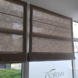 Volodymyr Vorobets - Dwie brązowe rolety rzymskie zamontowane w oknie. Widoczna faktura materiału i mechanizm podnoszenia. W tle zieleń za oknem i logo firmy na szybie.