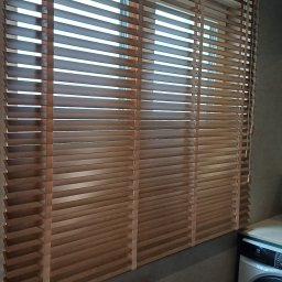 Volodymyr Vorobets - Zamknięte żaluzje drewniane w łazience, widoczne sznury do regulacji. Częściowo widoczna wanna i pralka. Naturalne światło dzienne.