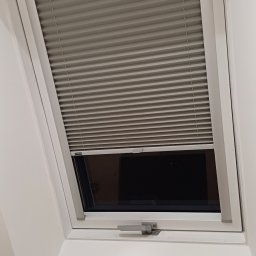 Volodymyr Vorobets - Szara plisa okienna zamontowana w oknie dachowym, widoczny mechanizm regulacji, biała rama okna, wnętrze domu z widocznym fragmentem ściany.