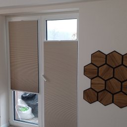 Volodymyr Vorobets - Beżowe plisy okienne, częściowo zasłonięte, na dwuskrzydłowym oknie PCV. Na ścianie dekoracja z drewnianych heksagonów.