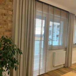 Volodymyr Vorobets - Beżowe żaluzje drewniane i zasłony w nowoczesnym wnętrzu z ceglaną ścianą i rośliną doniczkową. Widok na okno z widokiem na zewnątrz.