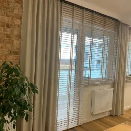 Beżowe żaluzje drewniane i zasłony w nowoczesnym wnętrzu z ceglaną ścianą i rośliną doniczkową. Widok na okno z widokiem na zewnątrz.