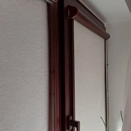 Volodymyr Vorobets - Jasne rolety wewnętrzne zamontowane w oknie o ciemnobrązowej ramie. Widoczny mechanizm łańcuszkowy i uchwyt okna. W tle firanka i roślina doniczkowa.