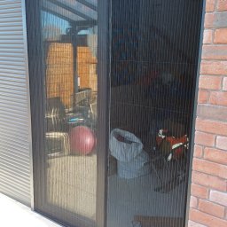 Volodymyr Vorobets - Zamknięta, plisowana moskitiera w kolorze czarnym, zamontowana w drzwiach tarasowych. Widoczna ceglana ściana i fragment szarej elewacji. Na tarasie leży piłka.
