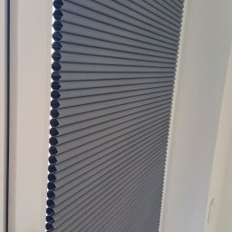 Volodymyr Vorobets - Szara plisa okienna zamontowana w białej ramie okiennej. Widoczny mechanizm sterujący. Plisa częściowo zasłonięta, tworzy regularne, poziome fałdy.