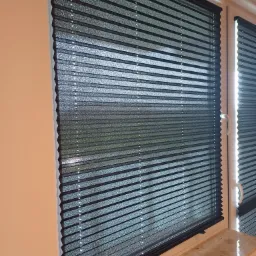 Elegancka, grafitowa plisa okienna zamontowana w oknie PCV. Widoczne delikatne załamania materiału i subtelne światło prześwitujące przez tkaninę.