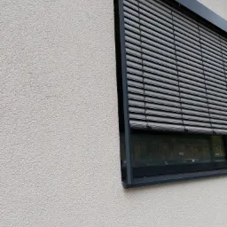 Zamknięte, szare żaluzje fasadowe zamontowane w oknie budynku z białą, chropowatą elewacją.