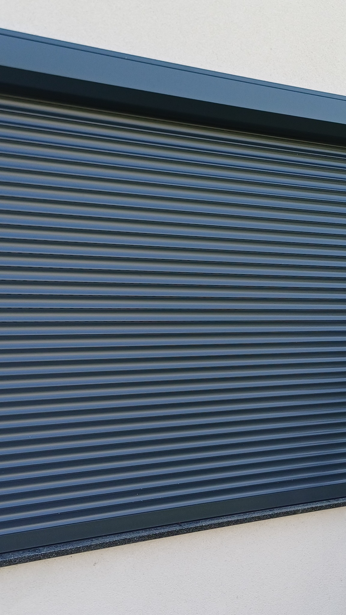 Zamknięta, grafitowa roleta zewnętrzna na elewacji budynku. Widoczne poziome lamele i kaseta rolety.