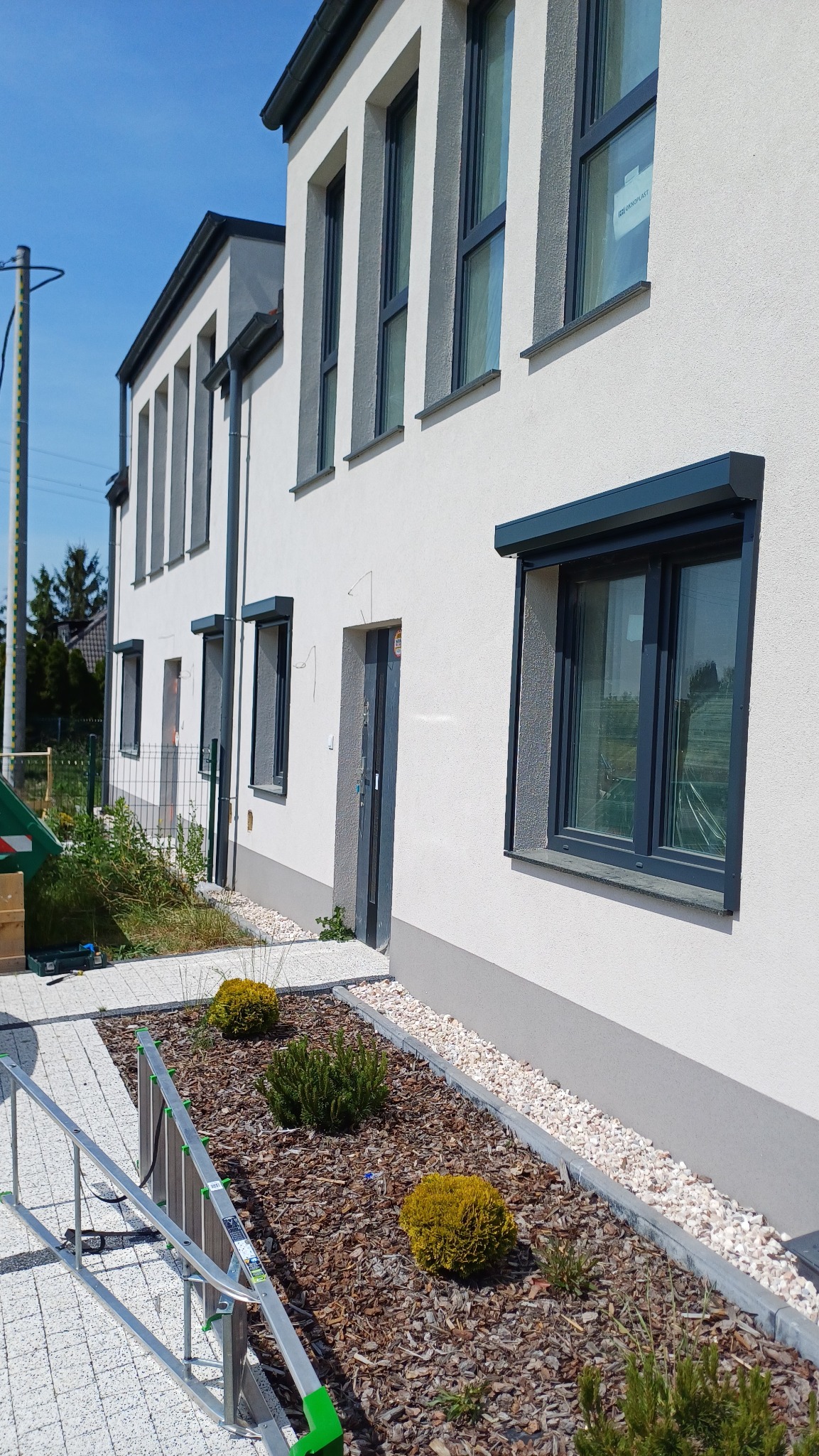 Elewacja budynku z zamontowanymi roletami zewnętrznymi w kolorze antracytowym. Widoczny fragment drabiny i rabaty z korą.