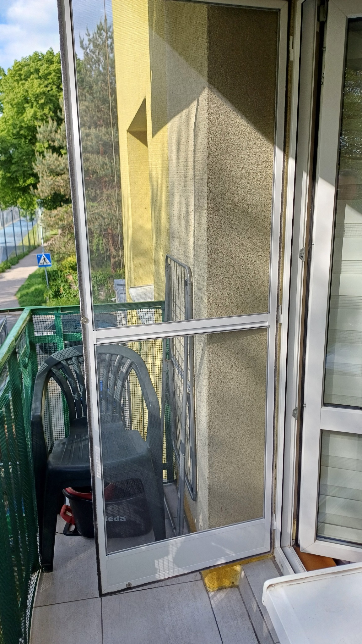 Zamontowana moskitiera ramkowa w drzwiach balkonowych, widok z balkonu na zieleń i ulicę. Dwa krzesła i suszarka na pranie na balkonie.