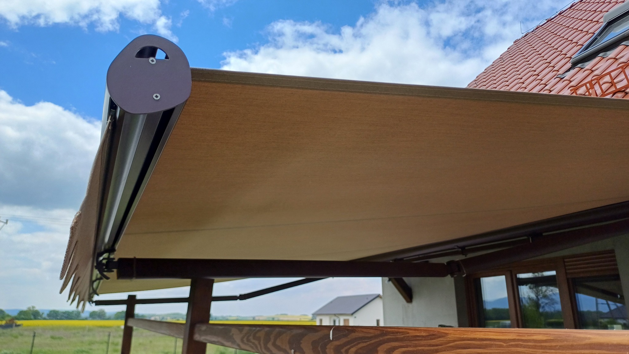 Beżowa markiza tarasowa z falbaną, zamontowana przy drewnianej konstrukcji. W tle widoczne pole rzepaku, dom i błękitne niebo z chmurami.