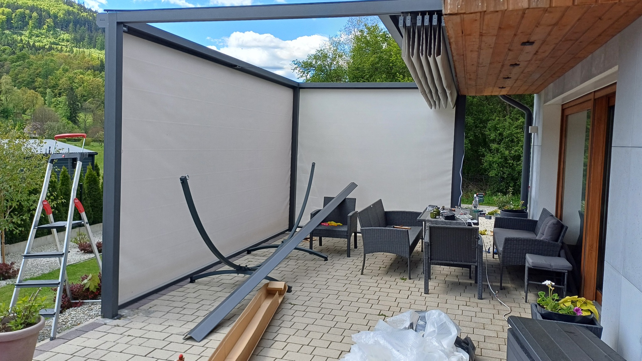 Montaż pergoli tarasowej z rolowanymi przesłonami bocznymi i zadaszeniem lamelowym. Widoczny drabina, narzędzia i elementy konstrukcji w trakcie instalacji.