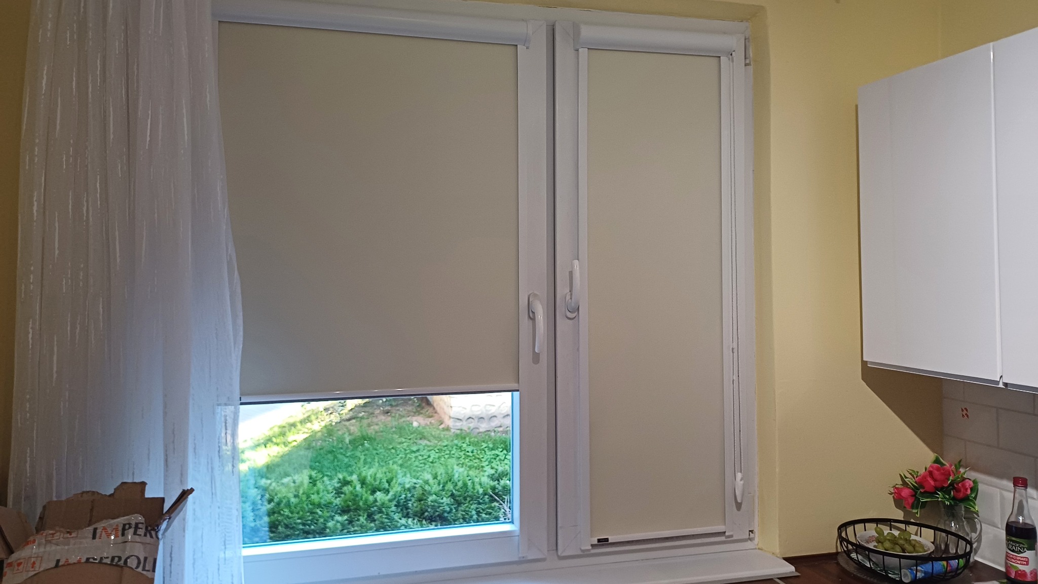 Beżowe rolety zwijane w oknach PCV, jedna częściowo opuszczona, z widokiem na zieleń. Obok firanka i fragment blatu kuchennego z dekoracjami.