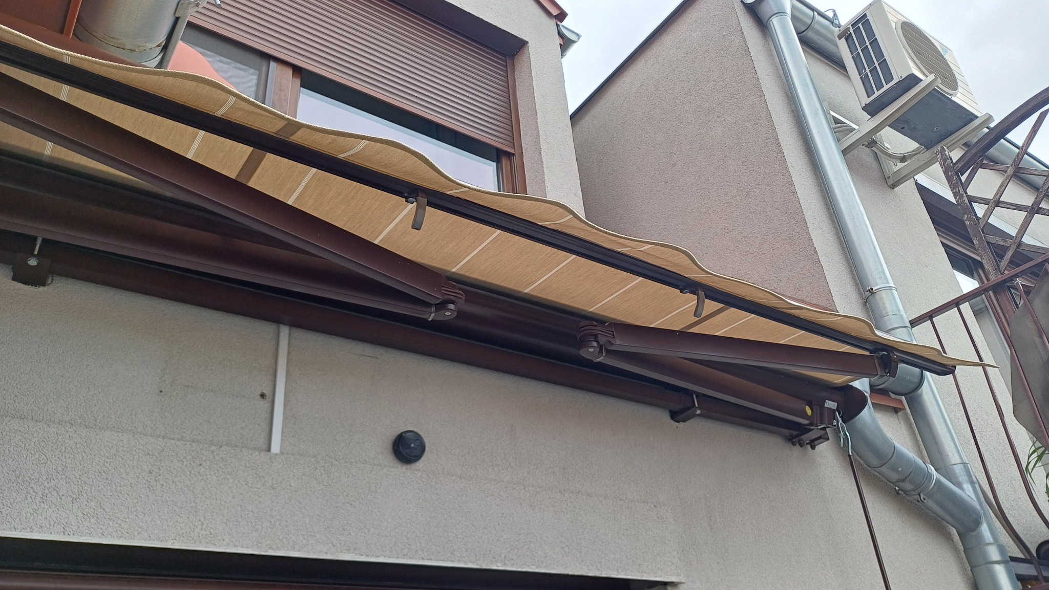 Beżowa markiza tarasowa z brązową konstrukcją zamontowana nad oknem z roletą zewnętrzną. Widoczny fragment rynny i klimatyzator na elewacji budynku.