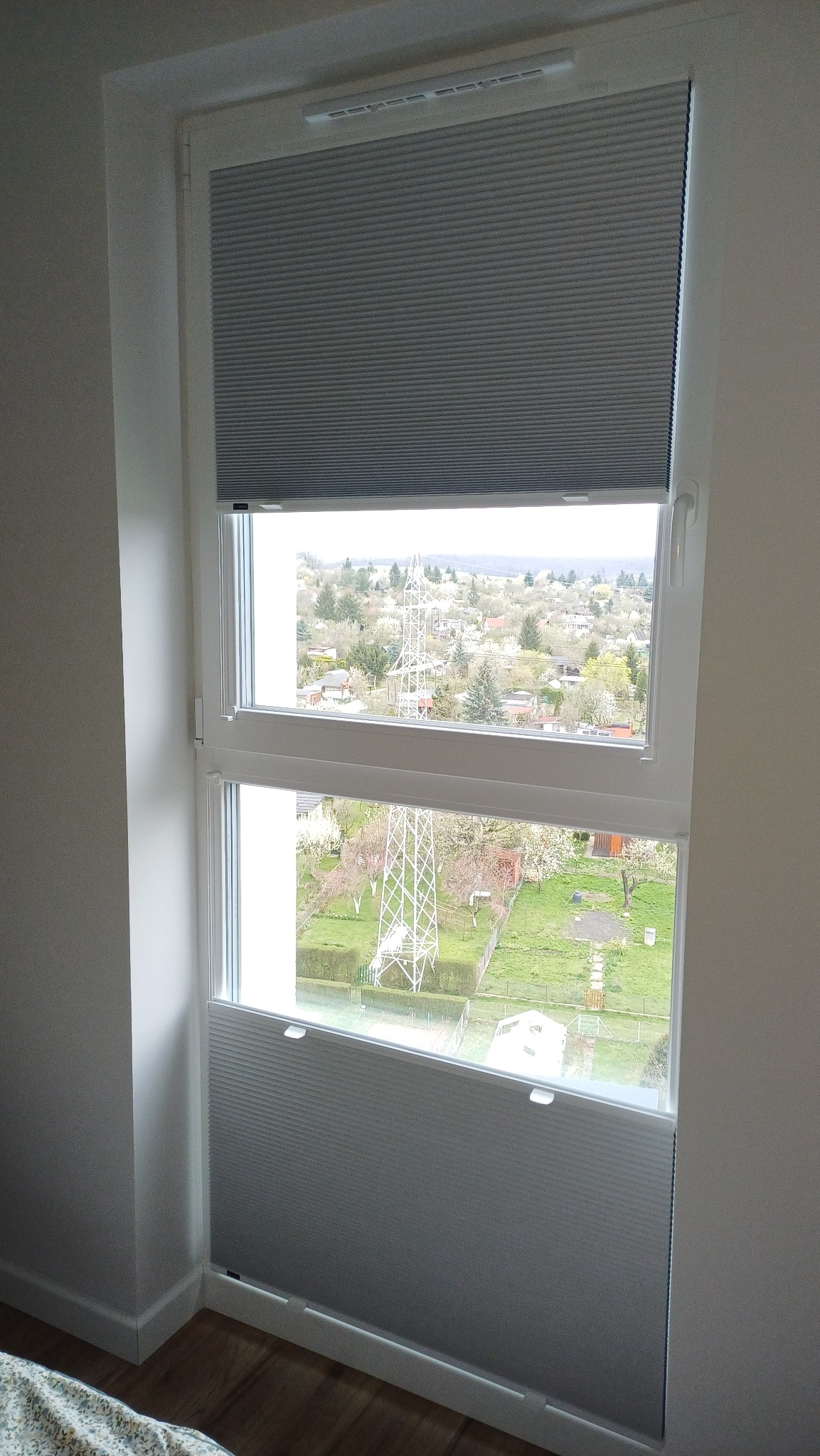 Szara, podwójna plisa okienna w Świdnicy, zamontowana w oknie z widokiem na okolicę. Górna i dolna część okna zasłonięta, środkowa odsłonięta.