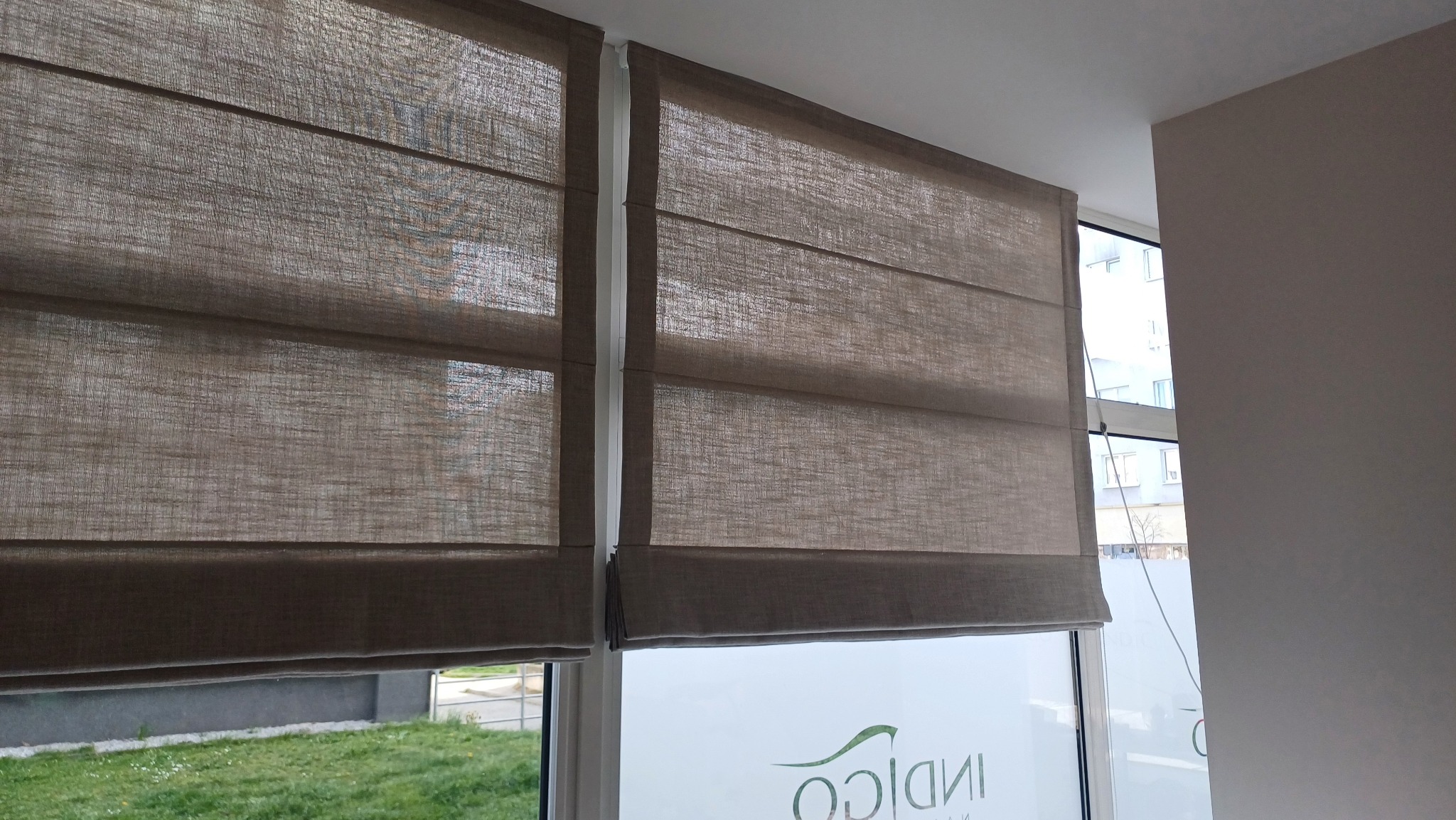 Dwie brązowe rolety rzymskie zamontowane w oknie. Widoczna faktura materiału i mechanizm podnoszenia. W tle zieleń za oknem i logo firmy na szybie.
