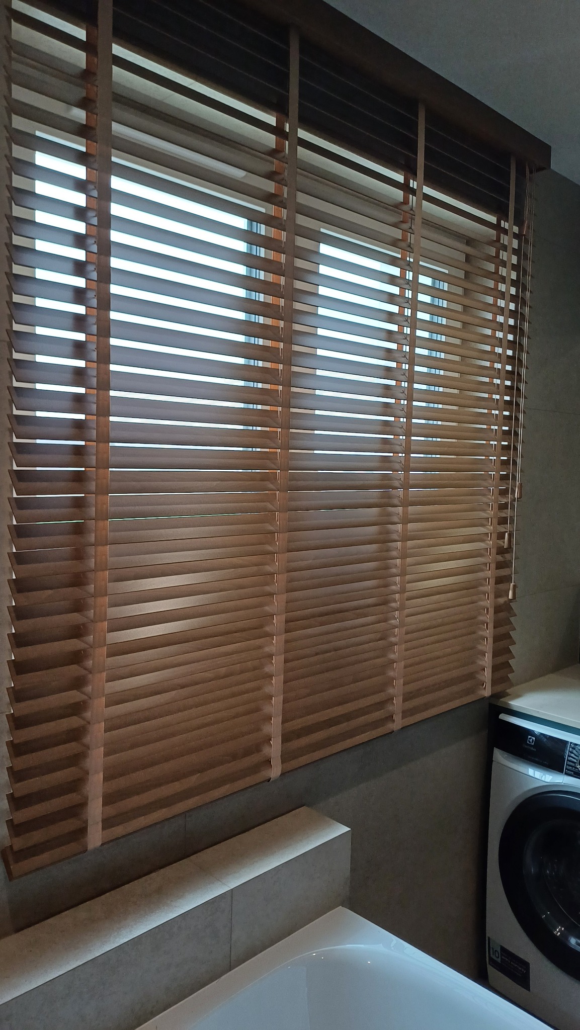 Zamknięte żaluzje drewniane w łazience, widoczne sznury do regulacji. Częściowo widoczna wanna i pralka. Naturalne światło dzienne.