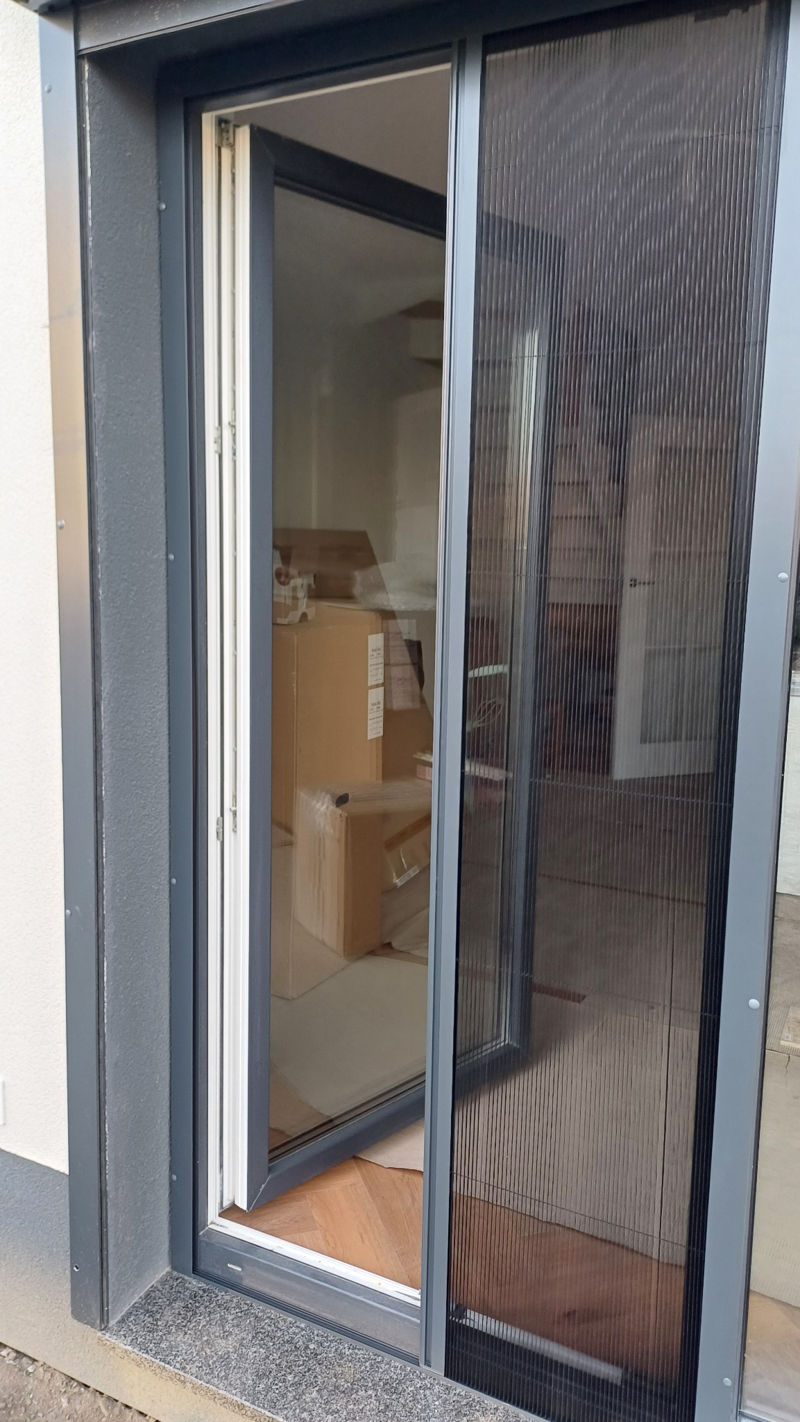 Rozsunięta moskitiera plisowana w grafitowej ramie, widok z zewnątrz na skrzydło okienne i wnętrze z kartonami. Ochrona przed owadami w nowoczesnym wydaniu.