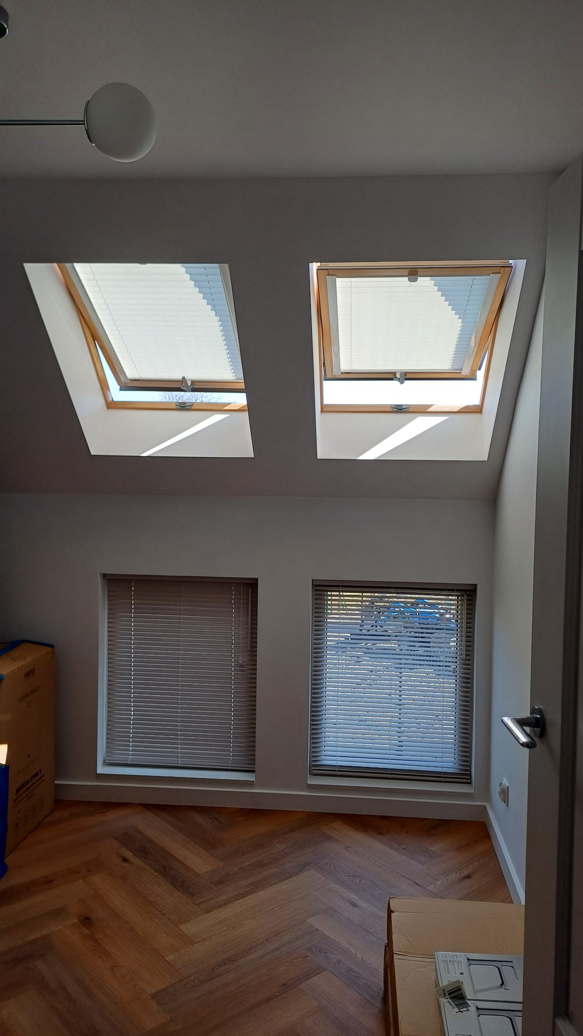 Dwa okna dachowe z plisami i dwa standardowe z żaluzjami drewnianymi w jasnym wnętrzu z parkietem w jodełkę. Plisy częściowo otwarte, żaluzje zamknięte.