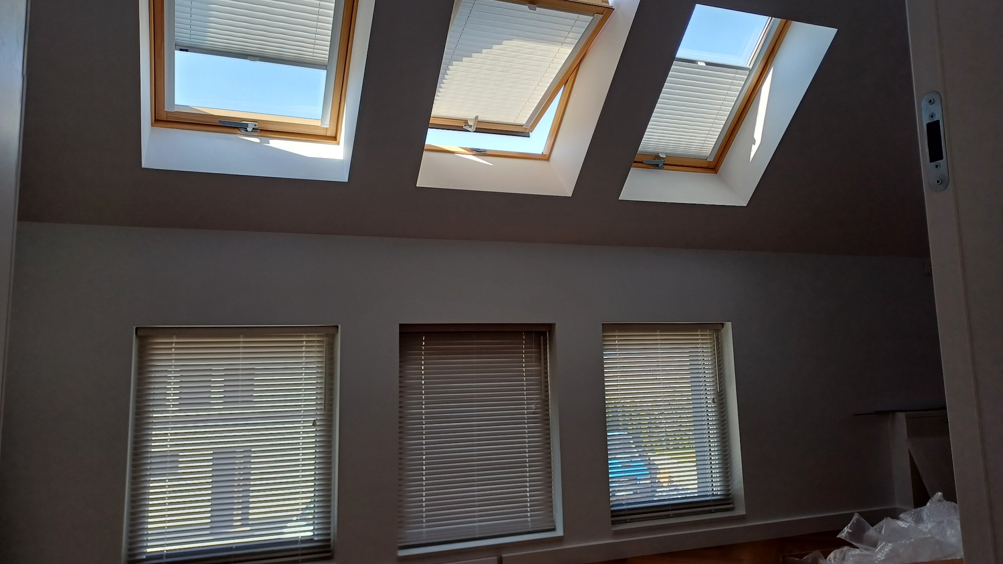 Trzy okna dachowe z plisowanymi roletami w różnym stopniu otwarcia, uzupełnione trzema oknami pionowymi z żaluzjami w szarym pomieszczeniu.