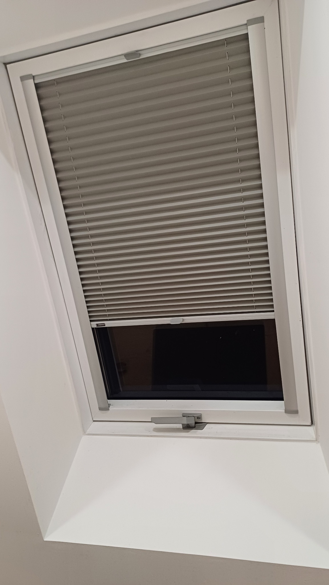 Szara plisa okienna zamontowana w oknie dachowym, widoczny mechanizm regulacji, biała rama okna, wnętrze domu z widocznym fragmentem ściany.