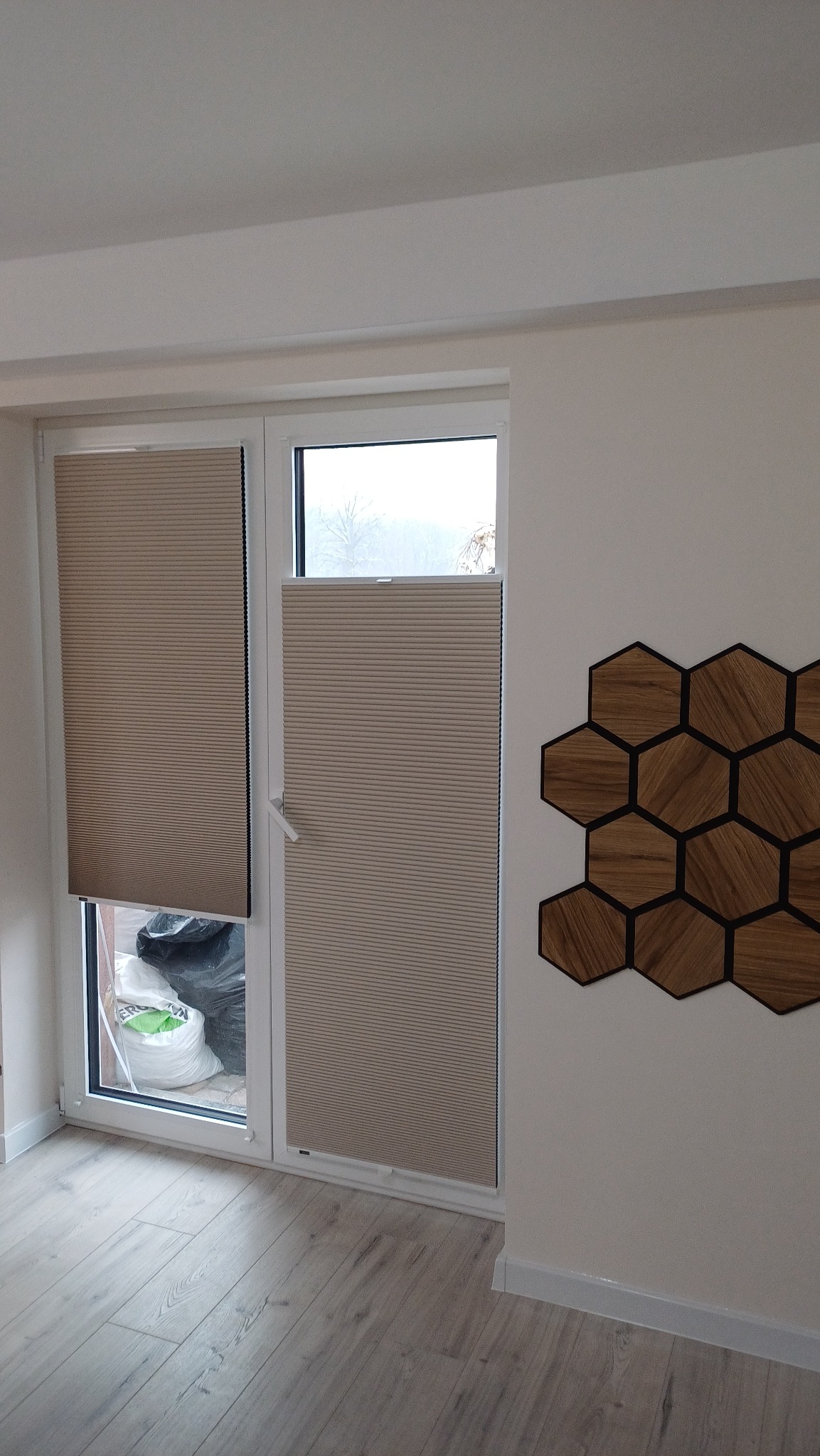 Beżowe plisy okienne, częściowo zasłonięte, na dwuskrzydłowym oknie PCV. Na ścianie dekoracja z drewnianych heksagonów.