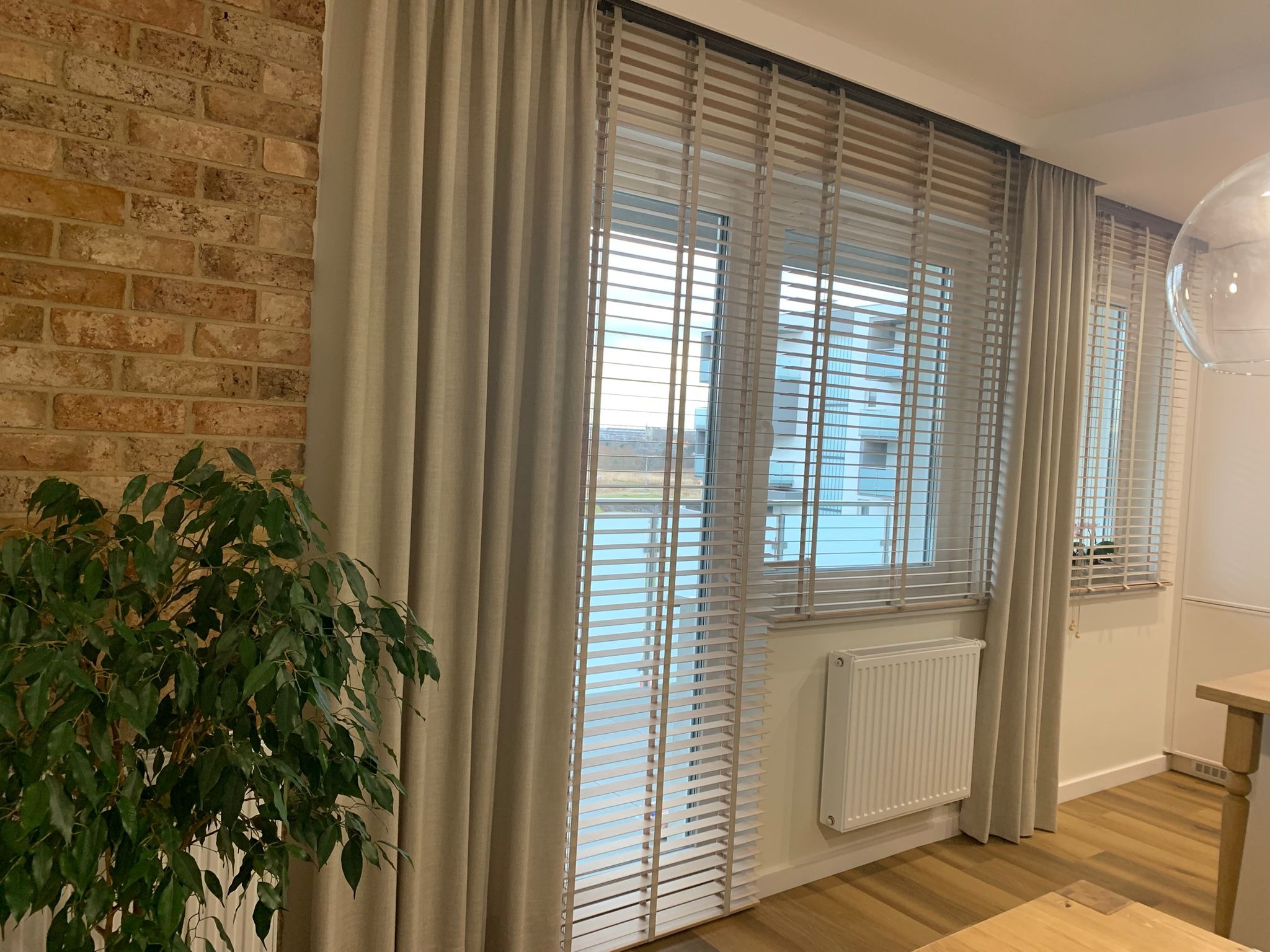 Beżowe żaluzje drewniane i zasłony w nowoczesnym wnętrzu z ceglaną ścianą i rośliną doniczkową. Widok na okno z widokiem na zewnątrz.