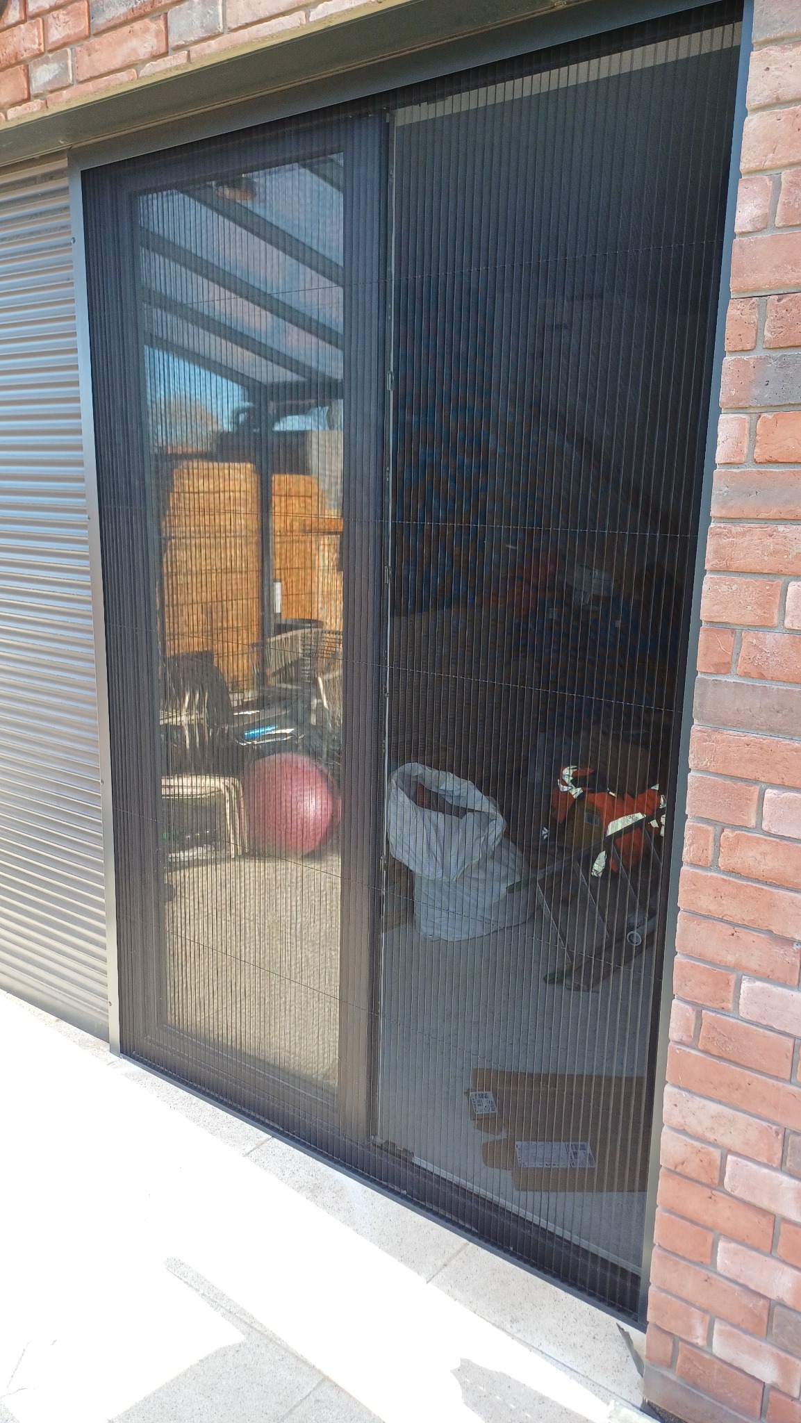 Zamknięta, plisowana moskitiera w kolorze czarnym, zamontowana w drzwiach tarasowych. Widoczna ceglana ściana i fragment szarej elewacji. Na tarasie leży piłka.