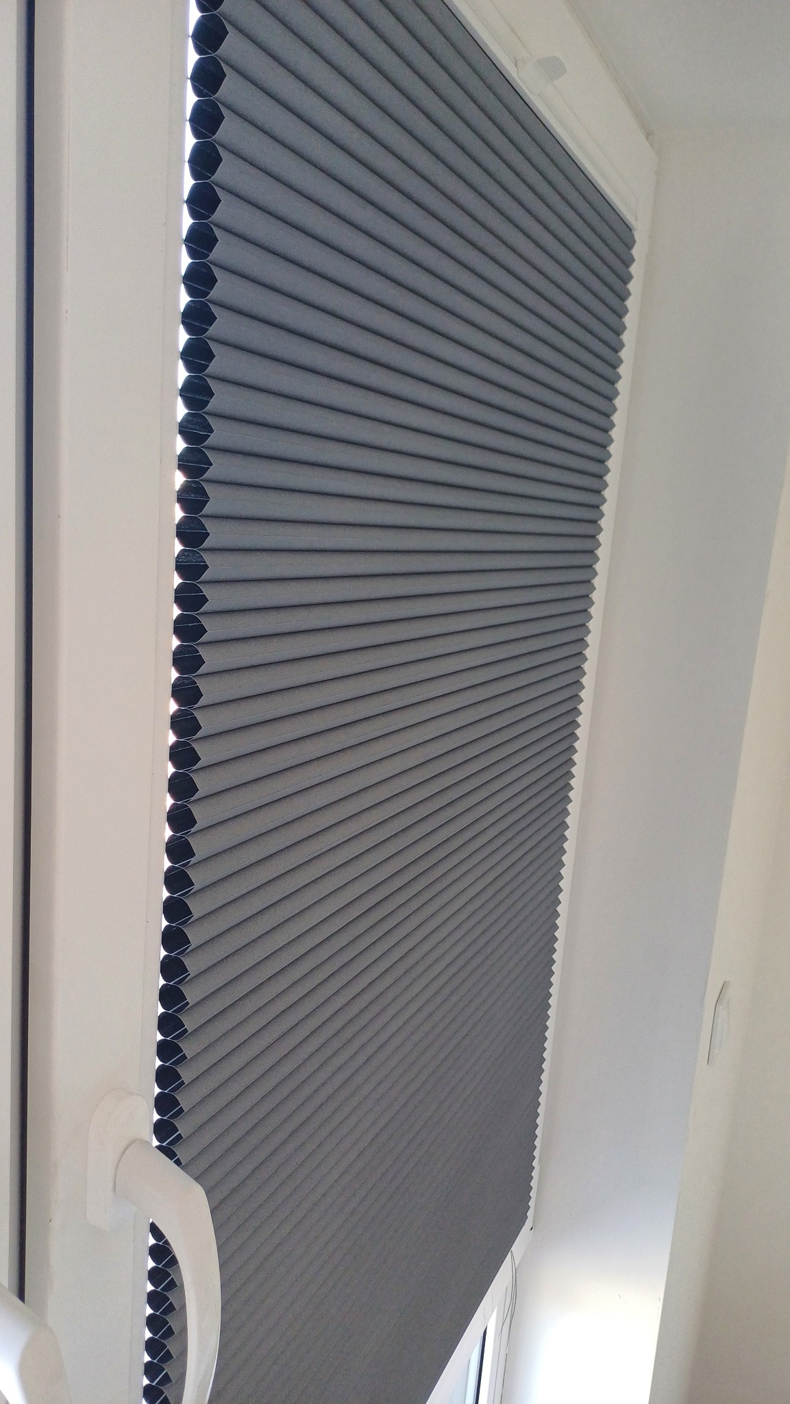 Szara plisa okienna zamontowana w białej ramie okiennej. Widoczny mechanizm sterujący. Plisa częściowo zasłonięta, tworzy regularne, poziome fałdy.
