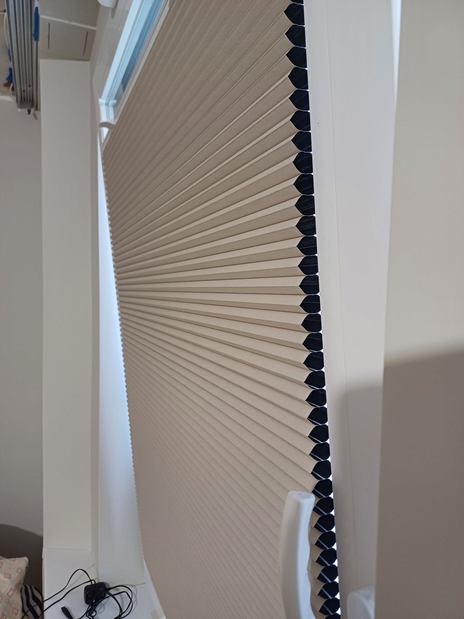 Beżowa plisa okienna zamontowana w białej ramie okiennej, widoczna z boku, z delikatnym światłem wpadającym przez okno. Widoczne przewody elektryczne w dolnej części.