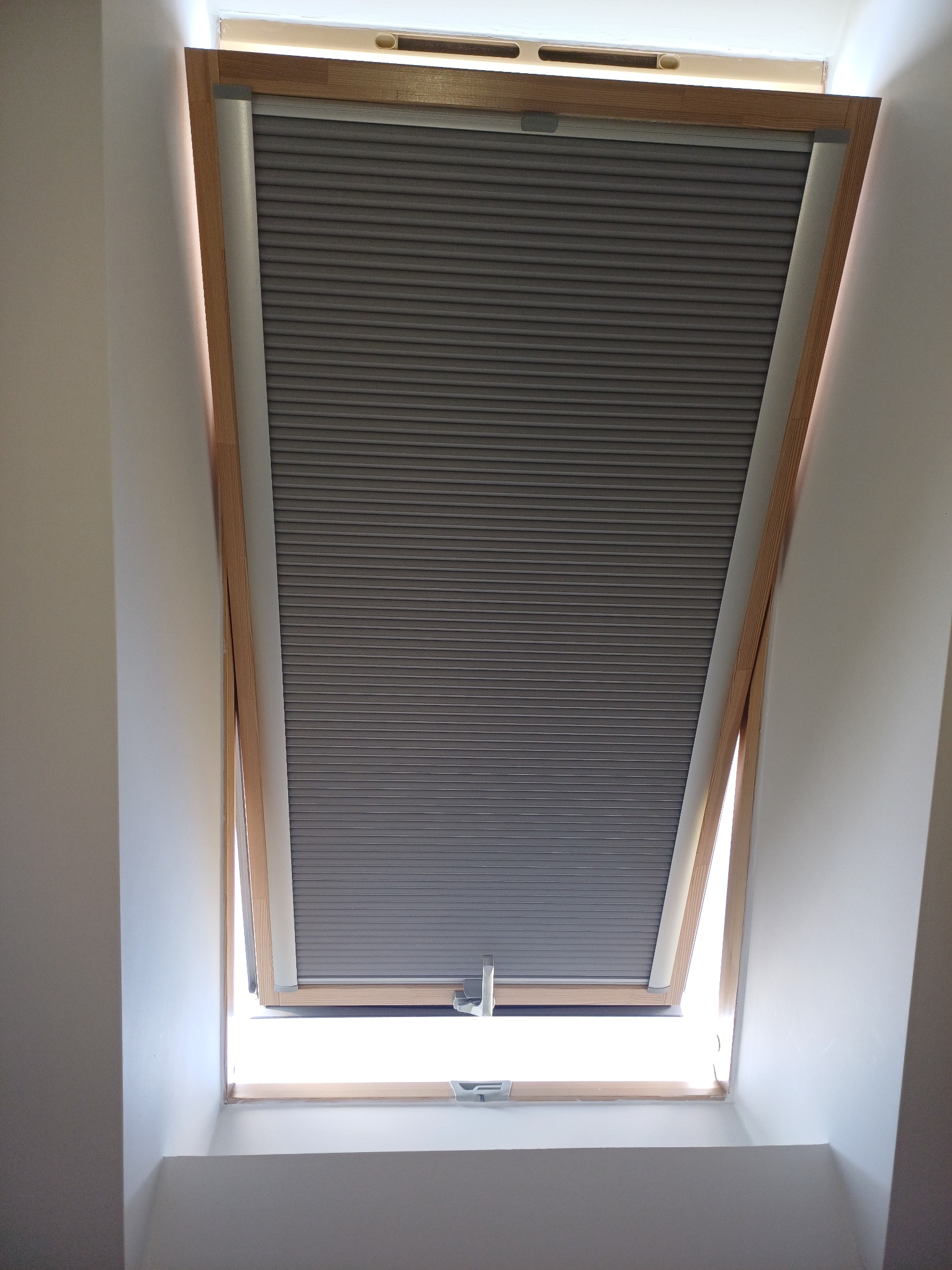 Szara plisa okienna zamontowana w oknie dachowym z drewnianą ramą. Widoczny mechanizm sterujący. Wpadające światło dzienne.