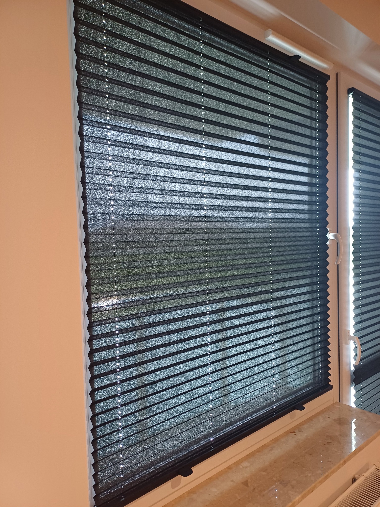 Elegancka, grafitowa plisa okienna zamontowana w oknie PCV. Widoczne delikatne załamania materiału i subtelne światło prześwitujące przez tkaninę.