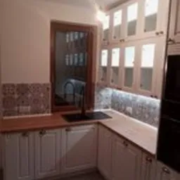 Kuchnia w stylu rustykalnym w Szadku. Białe szafki z drewnianymi blatami i przeszklonymi górnymi frontami, czarny kran i okno w drewnianej ramie. Nad blatem dekoracyjna mozaika.