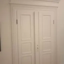 Białe, dwuskrzydłowe drzwi wewnętrzne z dekoracyjną ramą i panelami, widoczne zawiasy i klamka, sfotografowane z lekkiego dołu pod kątem.