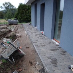 Blumen - Ułożona ścieżka z szarej kostki brukowej przy ścianie domu, z widocznym brudnym wózkiem budowlanym i paletą kostki w tle, teren przygotowany pod dalsze prace.