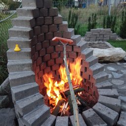 Blumen - Ognisko z cegieł i betonowych bloczków, z płomieniami i rożnem z kiełbasą, w tle ogród i sprzęt budowlany.