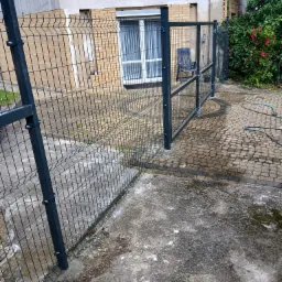 Nowoczesne ogrodzenie panelowe w kolorze grafitowym, oddzielające posesję od sąsiada. Widoczny fragment domu z oknem i fragment ogrodu z kostką brukową.