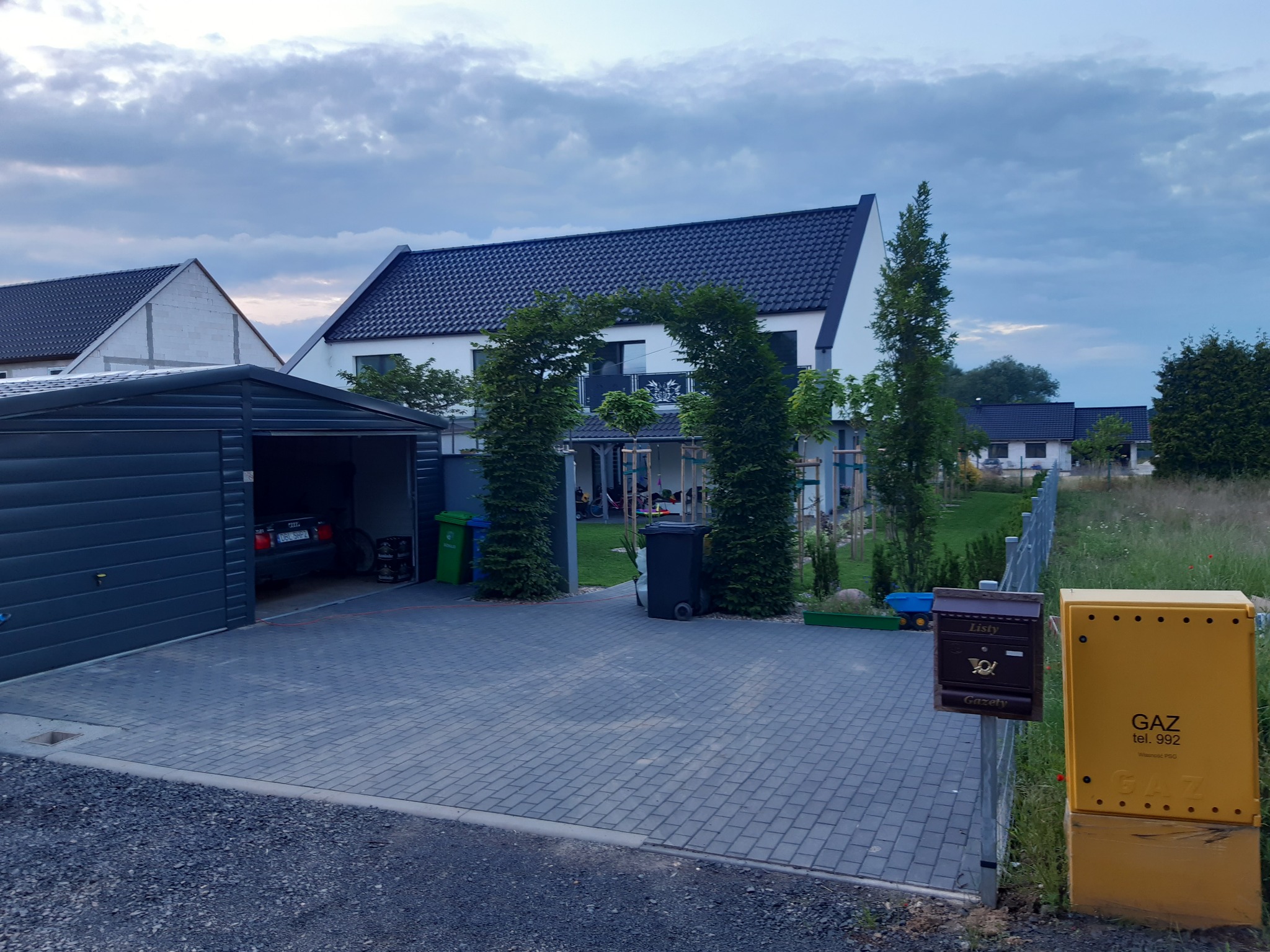 Ułożona kostka brukowa na podjeździe przed domem z garażem i metalową bramką z dwoma skrzynkami pocztowymi, widoczne również drzewa i krzewy.