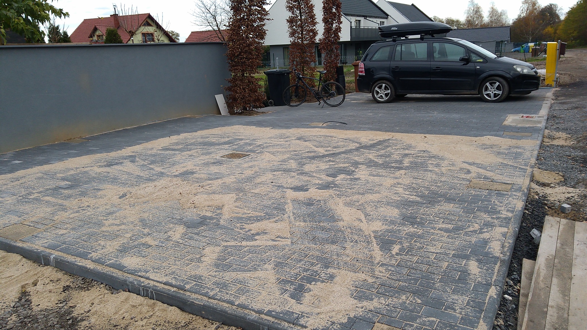 Niedawno ułożona szara kostka brukowa, zasypana piaskiem kwarcowym, z widocznym włazem rewizyjnym pośrodku, otoczona szarym murem i zielenią, z zaparkowanym czarnym kombi z bagażnikiem dachowym w tle.