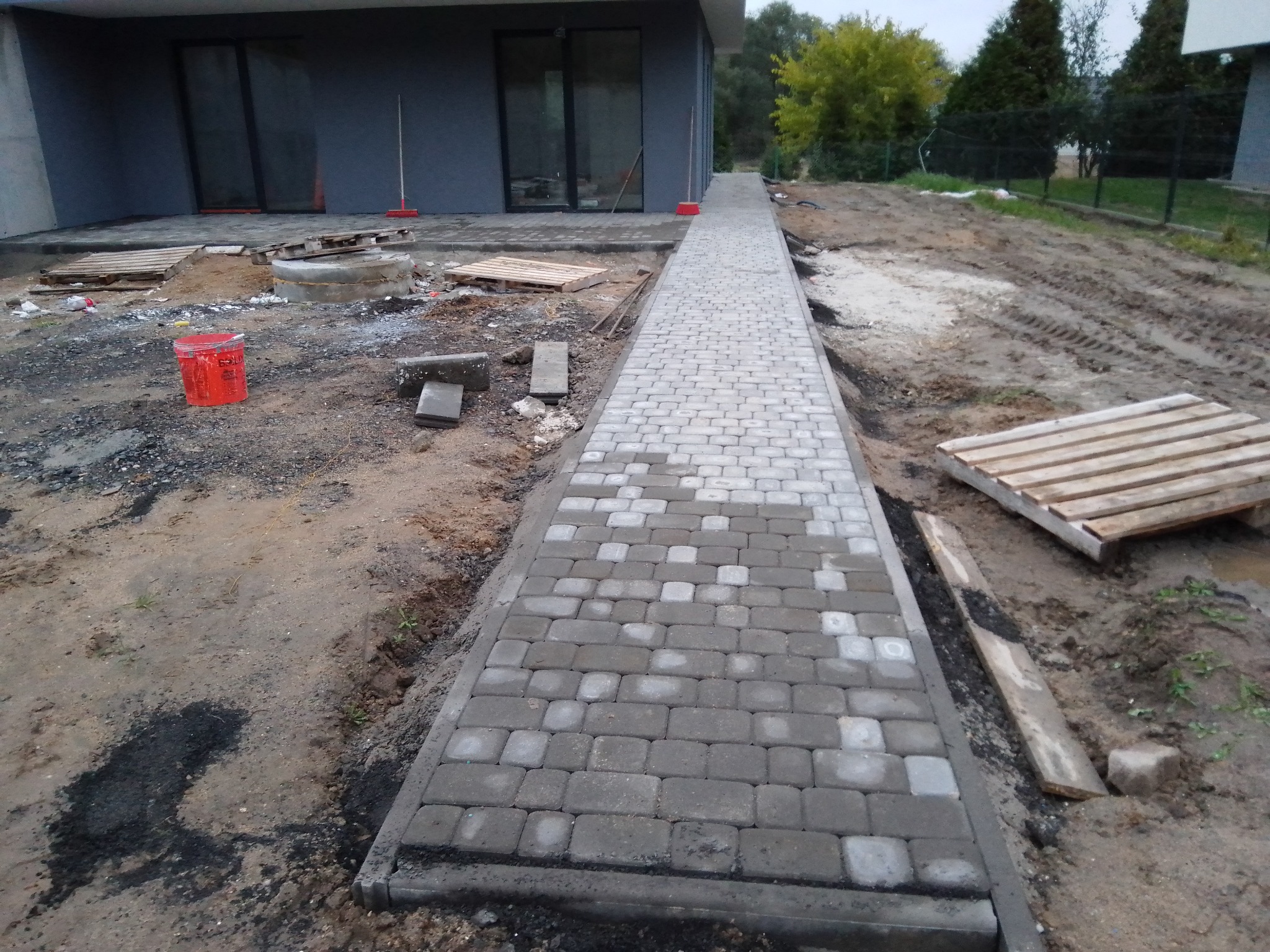 Częściowo ułożony chodnik z szarej kostki brukowej prowadzący do nowoczesnego domu z ciemną elewacją. Widoczne palety, wiadro i narzędzia budowlane sugerują trwające prace wykończeniowe.
