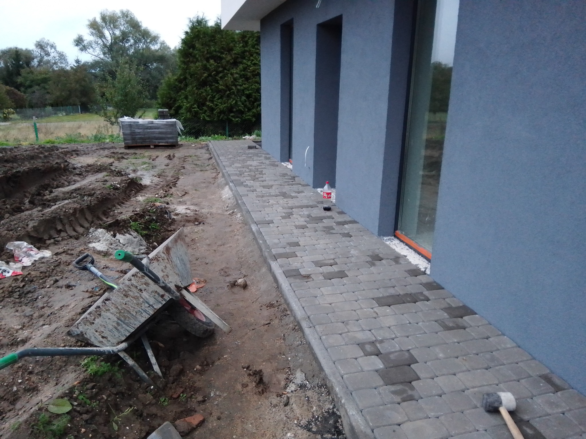 Ułożona ścieżka z szarej kostki brukowej przy ścianie domu, z widocznym brudnym wózkiem budowlanym i paletą kostki w tle, teren przygotowany pod dalsze prace.